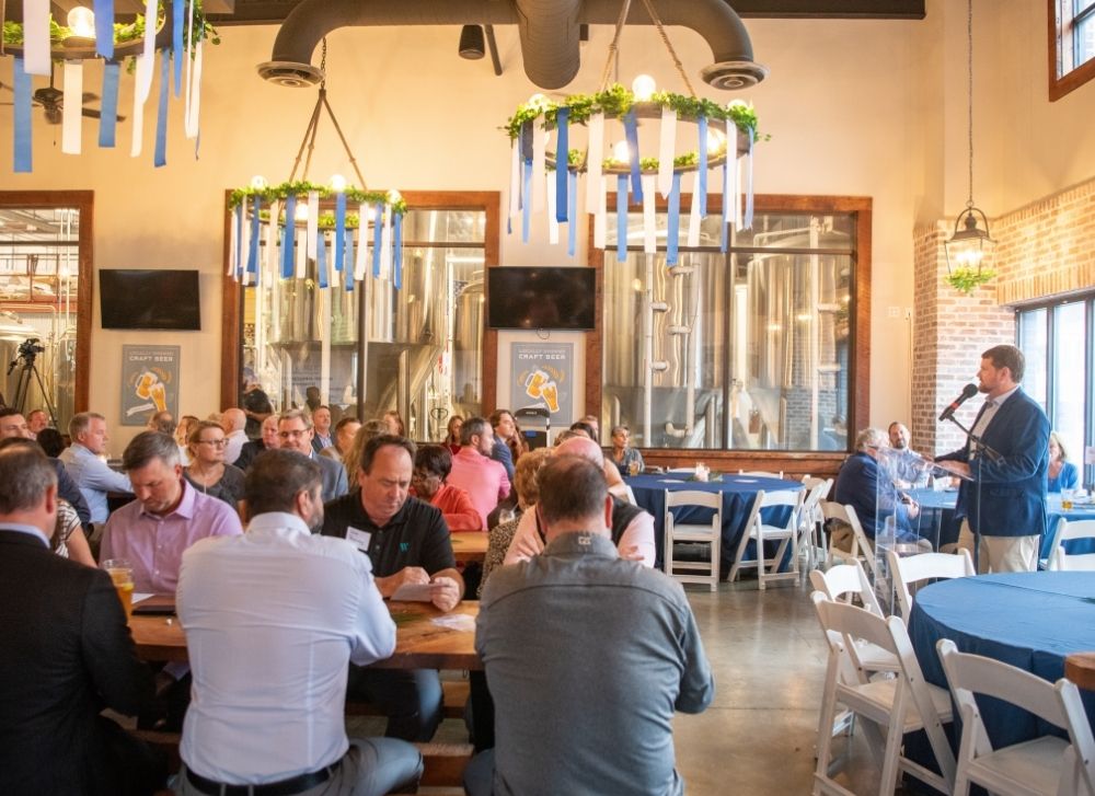 10 A group of people sit at tables in a brewery event space, listening to a man speaking at a podium. Blue ribbons and greenery hang from the ceiling as decorations.