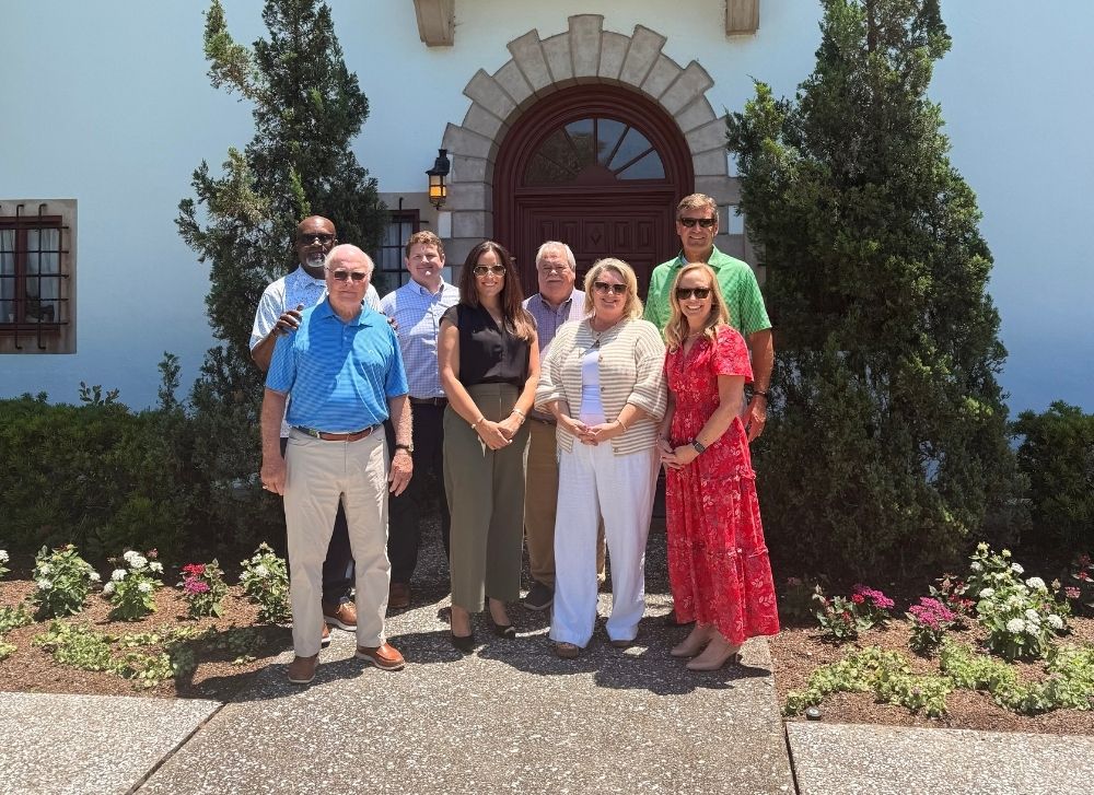 Eight adults stand in front of a building entrance, posing for a group photo on a sunny day. There are trees and flowers around the walkway.