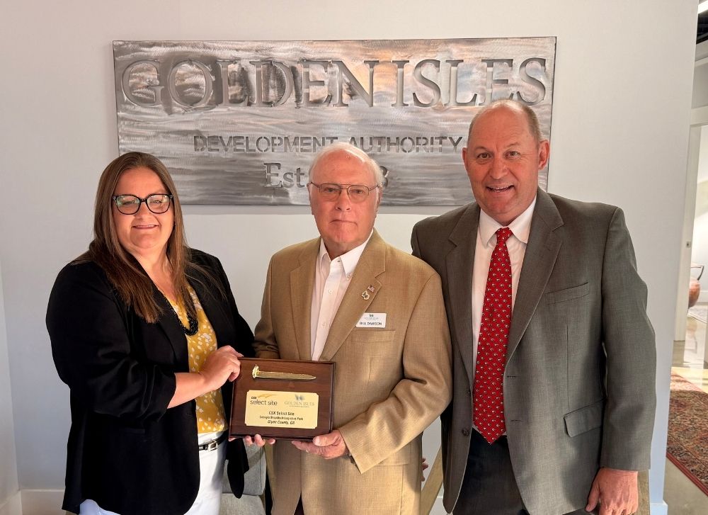 13 Three people in business attire stand together indoors; the man in the center holds a wooden plaque. A sign behind them reads Golden Isles Development Authority..