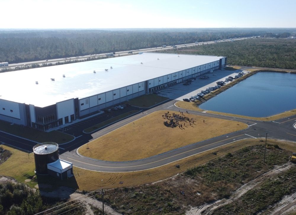 14 Aerial view of a large warehouse building with adjacent parking lot, small pond, and landscaped area, surrounded by forested land.