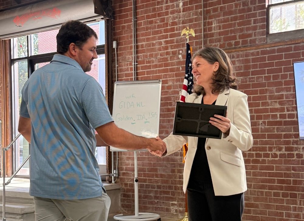15 A woman in a white blazer hands a plaque to a man in a blue shirt while they shake hands in front of a whiteboard and American flag in a brick-walled room.