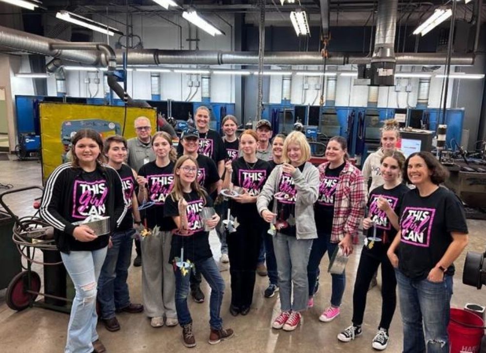 16 A group of people, mostly young women, stand in a workshop holding small trophies. Many wear black shirts reading THIS GIRL CAN. Industrial equipment is visible in the background.