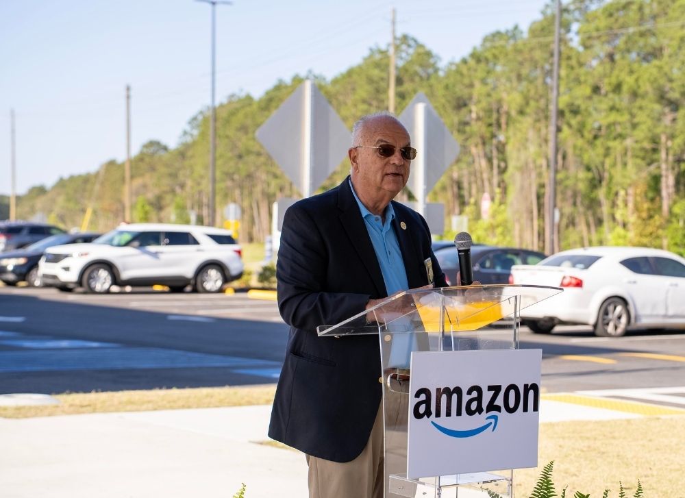 20 A man in a suit and sunglasses speaks at a podium with an Amazon sign outdoors, with parked cars and trees in the background.