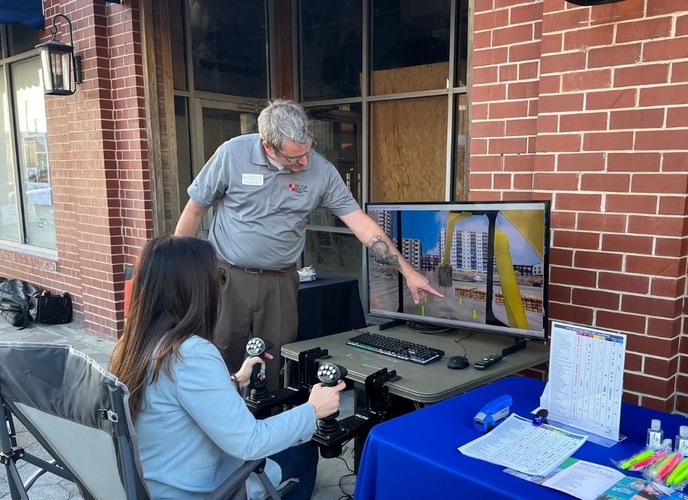 A man points at a screen displaying a construction simulation as a woman operates the simulator controls at an outdoor event booth.