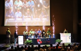 A group of people stands on stage at a GICCA National Signing Day event, with an audience watching, a large screen displaying the stage, and decorative lights in the background.