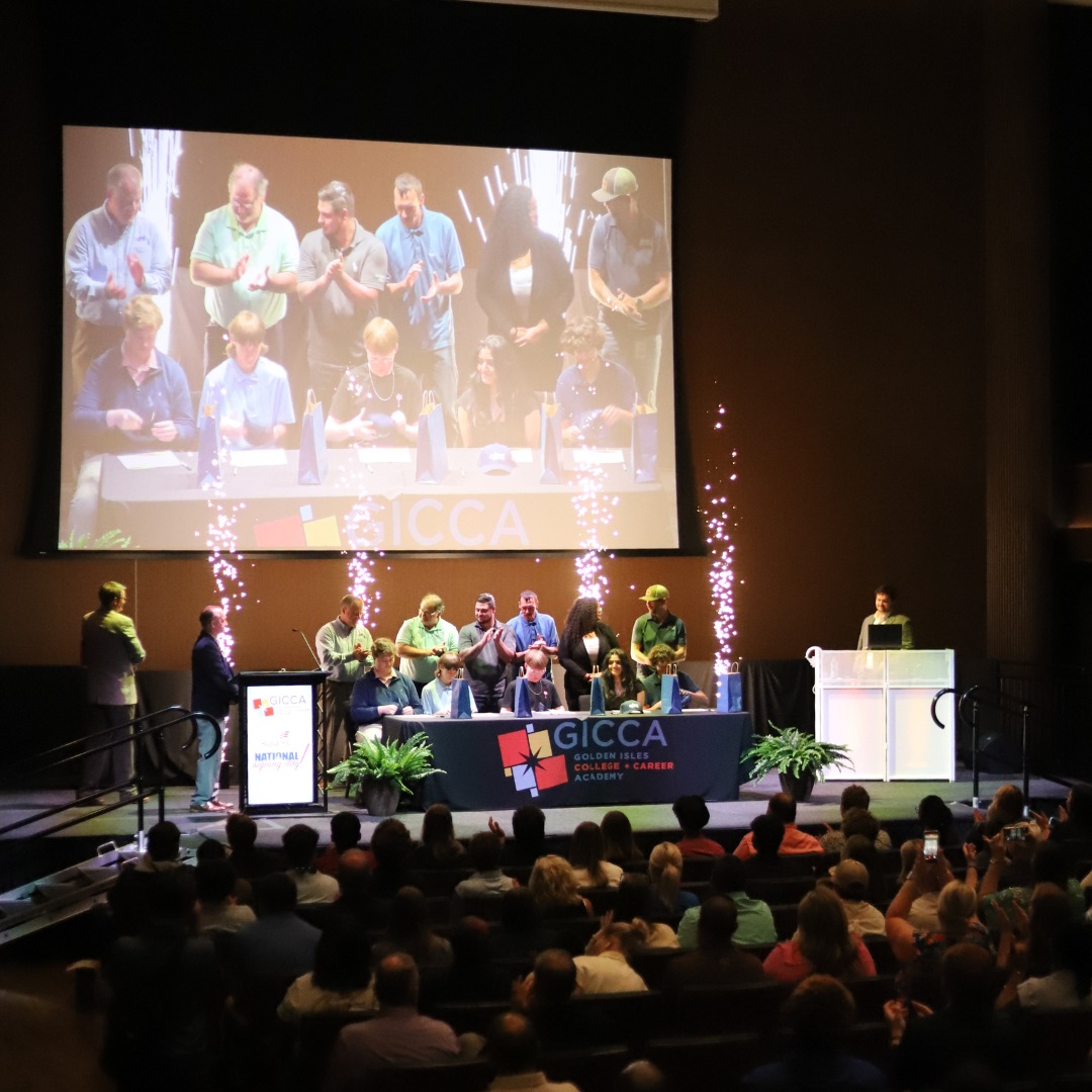 A group of people stands on stage at a GICCA National Signing Day event, with an audience watching, a large screen displaying the stage, and decorative lights in the background.