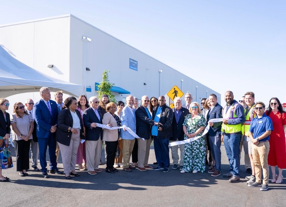 A group of people stands outside a large building during a ribbon cutting ceremony, holding a white ribbon and posing for a photo under clear skies.