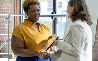 Two women shake hands as one presents an award plaque to the other indoors, celebrating GICCA National Signing Day in front of a large window and whiteboard.
