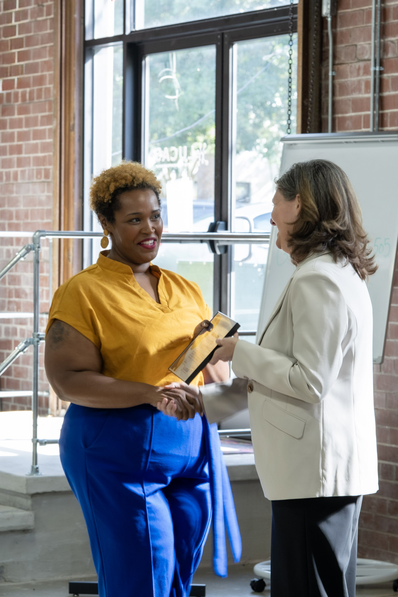 Two women shake hands as one presents an award plaque to the other indoors, celebrating GICCA National Signing Day in front of a large window and whiteboard.