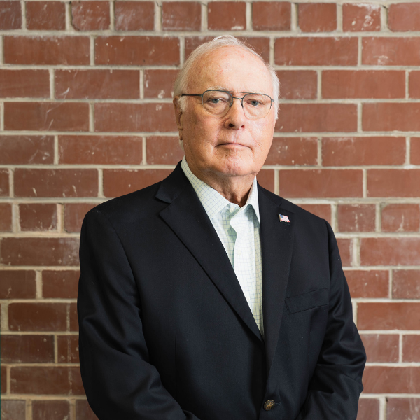 An older man in a dark suit jacket and glasses stands in front of a red brick wall, looking at the camera with a neutral expression.