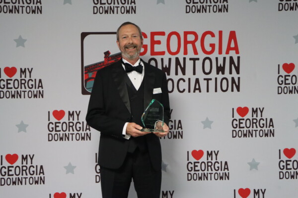 IMG_0160 Man in a tuxedo holding an award stands in front of a backdrop with Georgia Downtown Association and I Love My Georgia Downtown logos, celebrating the achievements of Elevate Glynn.