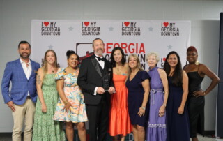 Nine people dressed in formal and semi-formal attire stand together indoors, smiling, with a My Georgia Downtown and Elevate Glynn step-and-repeat banner behind them. The man in the center holds an award.