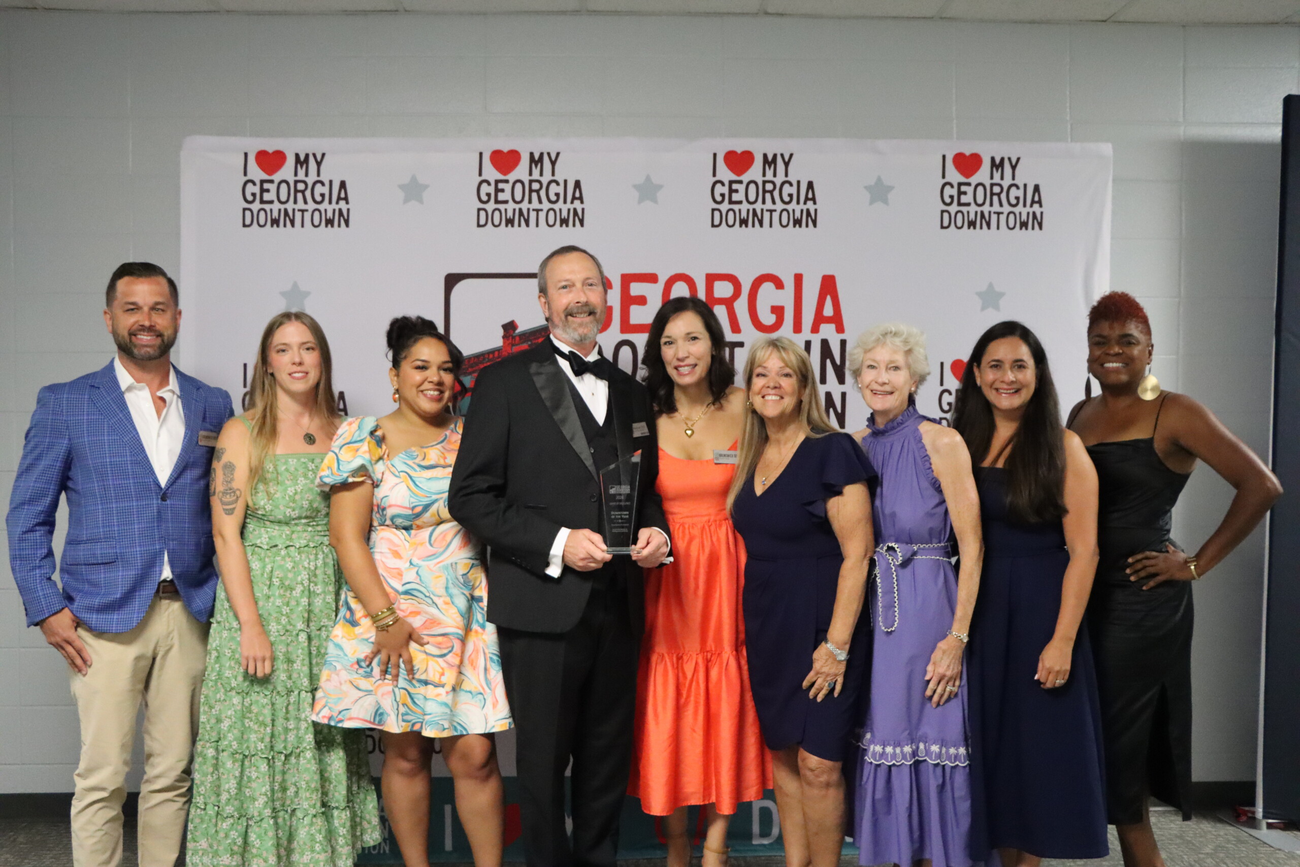 Nine people dressed in formal and semi-formal attire stand together indoors, smiling, with a My Georgia Downtown and Elevate Glynn step-and-repeat banner behind them. The man in the center holds an award.
