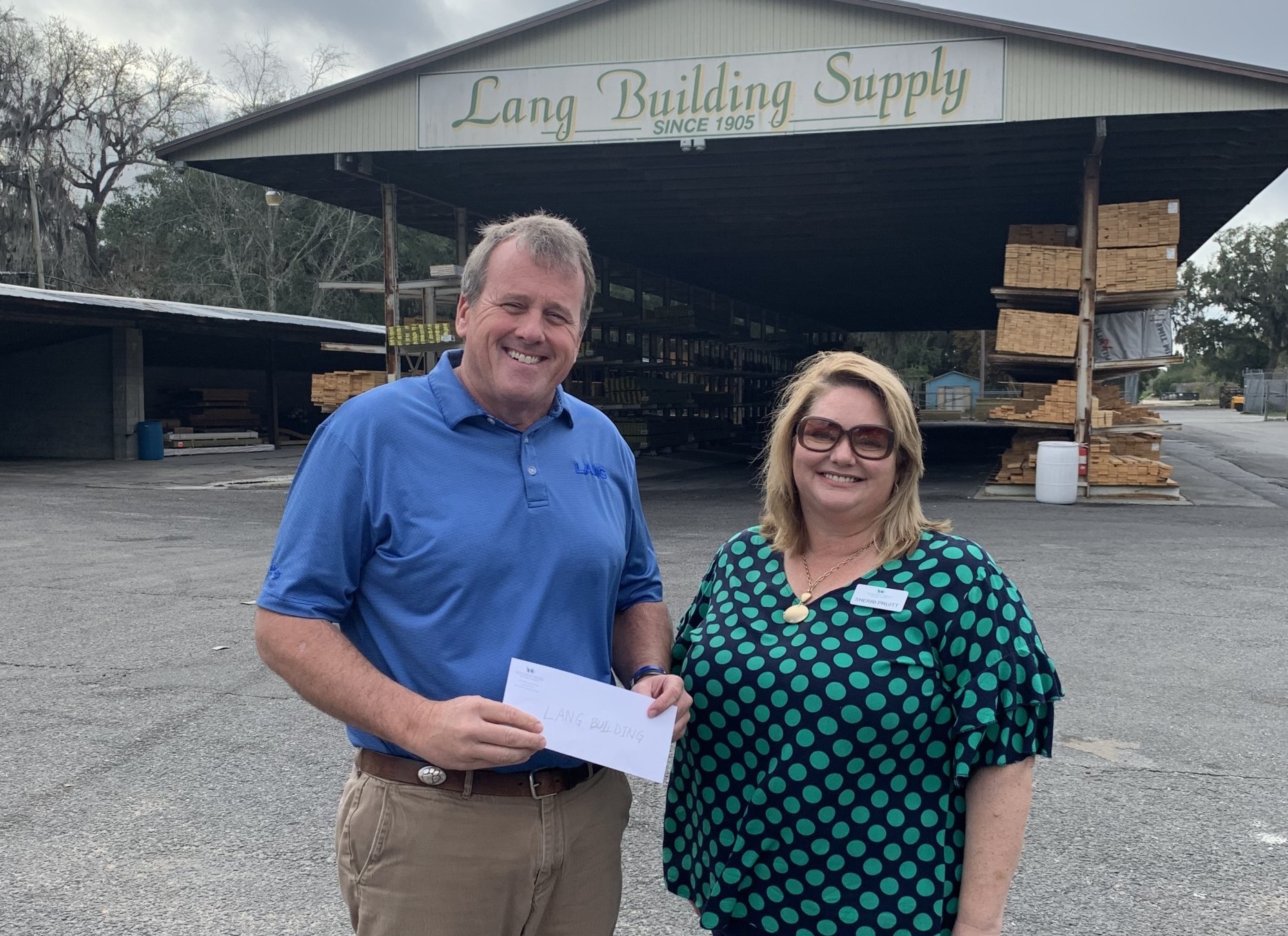LANG Storytelling_2 Two people stand in front of Lang Building Supply, smiling, as one holds a loan envelope. Stacks of lumber are visible under the building’s awning.
