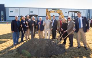 A group of people in business attire pose with shovels at a groundbreaking ceremony in front of construction equipment and a modern building, celebrating Downtown Brunswick 2026 Downtown of the Year.