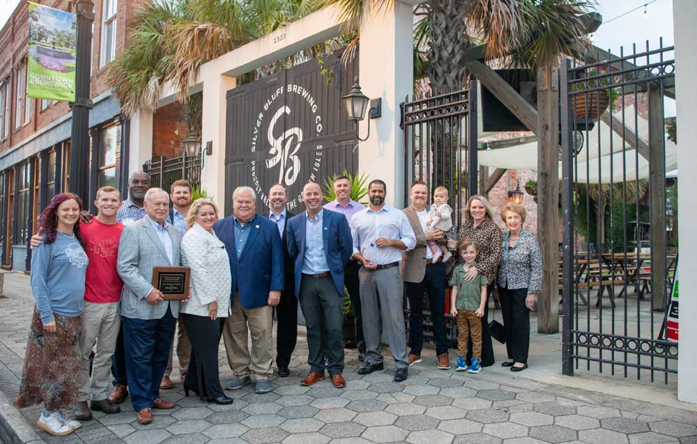 bluffbrewing A group of people poses for a photo outside a brewery entrance, with some holding plaques and others standing or sitting together, smiling at the camera—celebrating their contributions to the local economy.