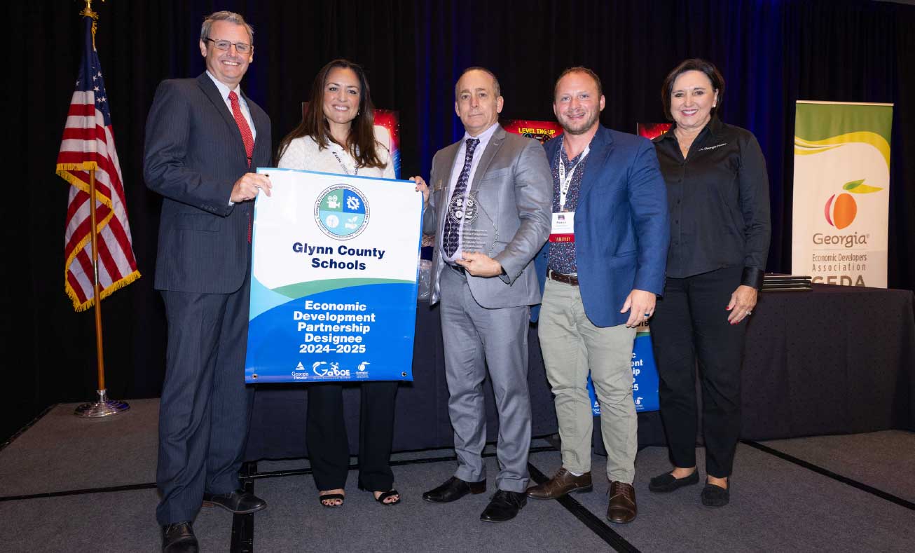 glynncountyschools Five people stand on stage holding a Glynn County Schools Economic Development Partnership banner, highlighting the area’s commitment to fostering business growth. Award plaques are shown, in front of a blue curtain and the U.S. and Georgia state flags.